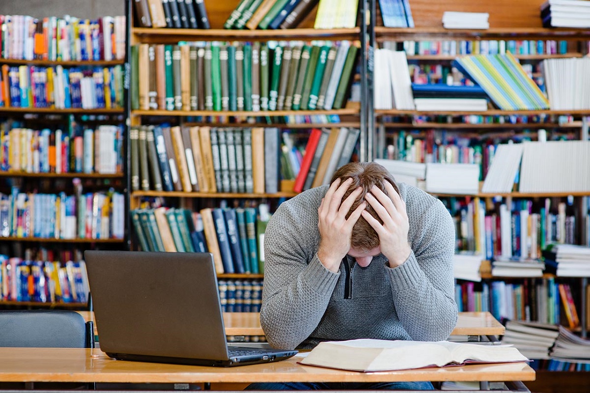 Joven estudiante con Ansiedad Académica visiblemente sobrecargado y ansioso, con las manos en la cabeza, sentado frente a libros y una computadora portátil en una biblioteca.